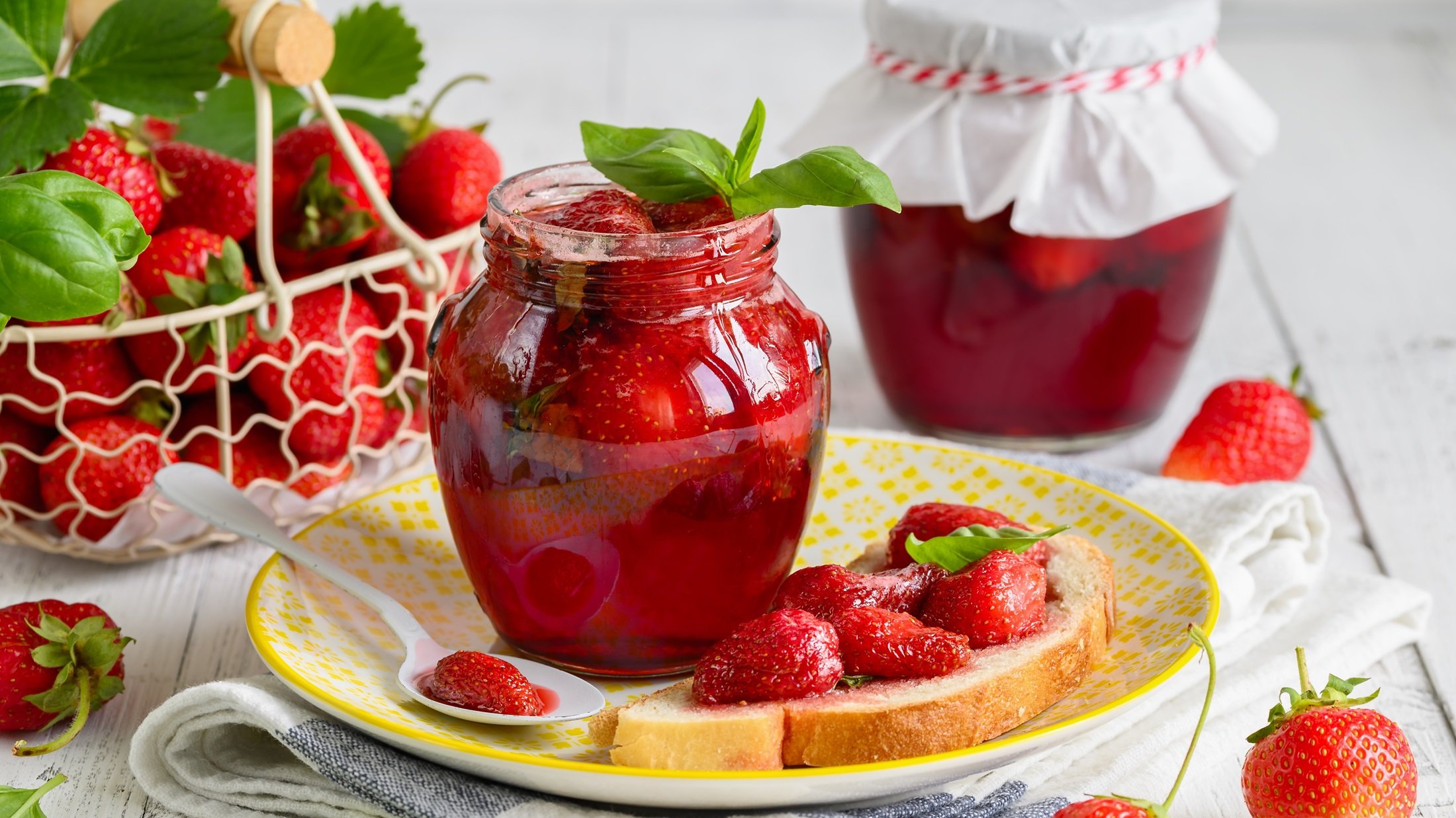 Frontal: Zwei Gläser Erdbeerkonfitüre mit Basilikum auf einem hellem Untergrund. Drum herum liegen frische Erdbeeren.