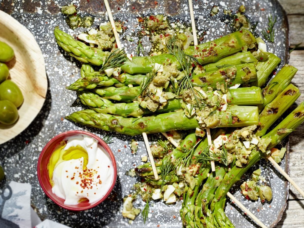 Grillspargel mit Kapern-Ei-Vinaigrette und Knobicreme auf einem grau-weißen-Teller angerichtet. Der Hintergrund ist hölzern.