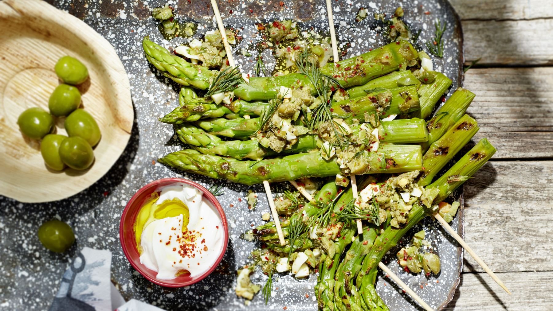Grillspargel mit Kapern-Ei-Vinaigrette und Knobicreme auf einem grau-weißen-Teller angerichtet. Der Hintergrund ist hölzern.