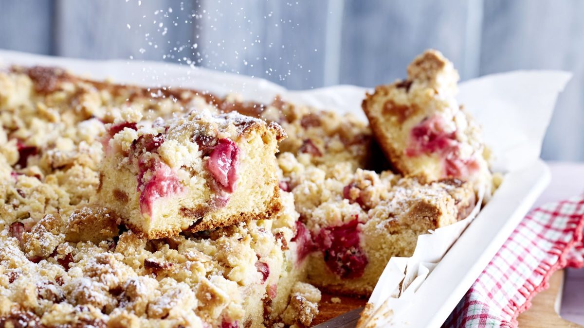 Landfräuleins Rhabarber-Streuselkuchen auf einem weißen Backblech auf einem rot-weiß-kariertem Tuch. Der Hintergrund ist grau.