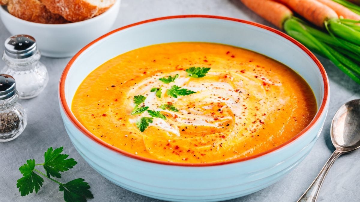 Draufsicht schräg; eine Kartoffel-Möhren-Suppe in einer runden Schüssel. daneben liegen ein Löffel und mehrere Möhren, beziehungswiese Karotten.