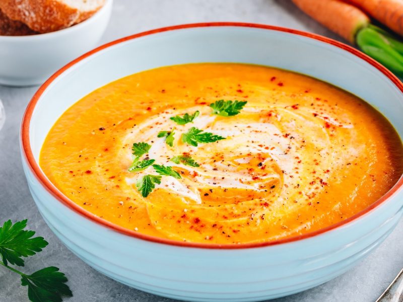 Draufsicht schräg; eine Kartoffel-Möhren-Suppe in einer runden Schüssel. daneben liegen ein Löffel und mehrere Möhren, beziehungswiese Karotten.