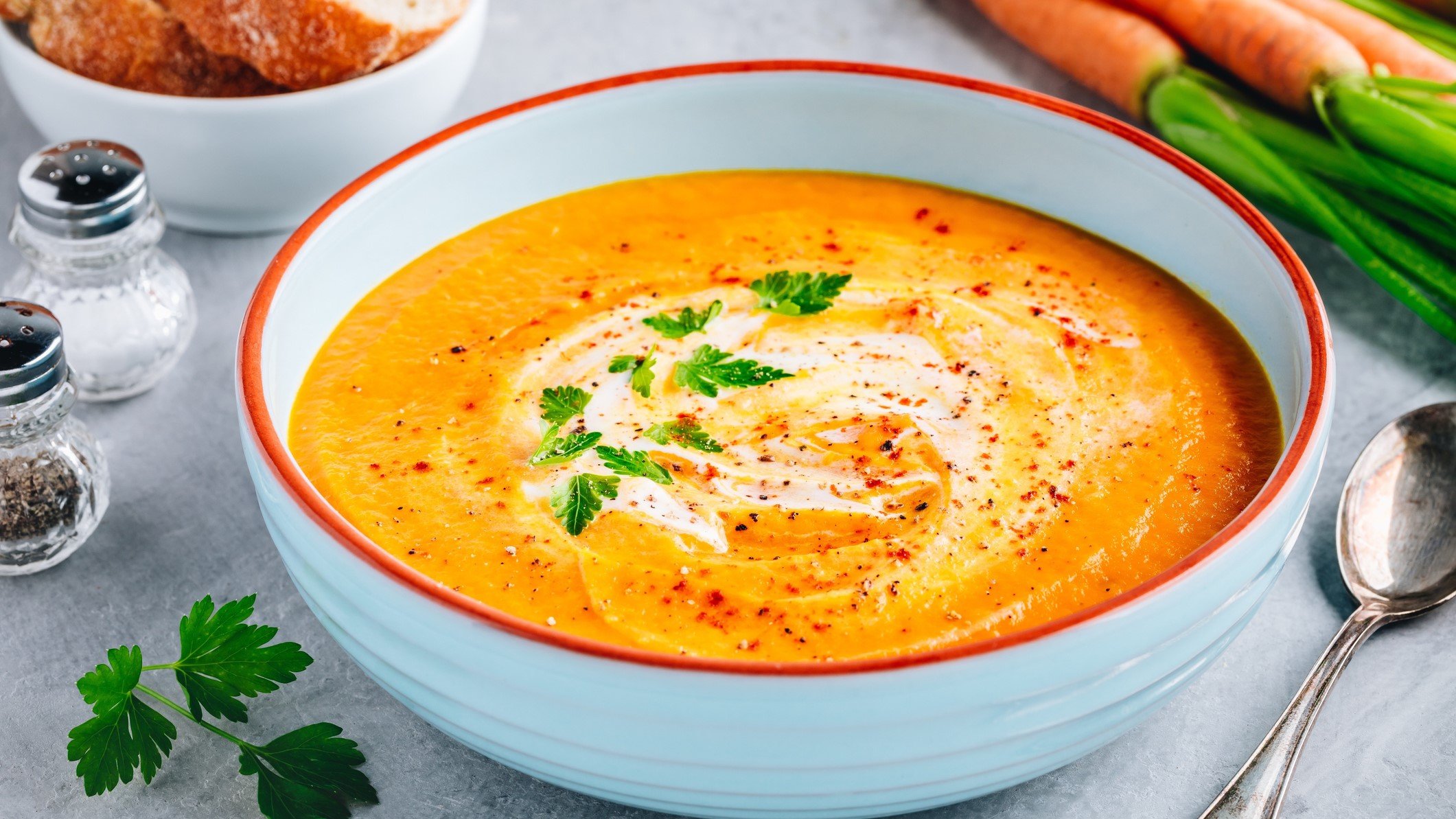 Draufsicht schräg; eine Kartoffel-Möhren-Suppe in einer runden Schüssel. daneben liegen ein Löffel und mehrere Möhren, beziehungswiese Karotten.