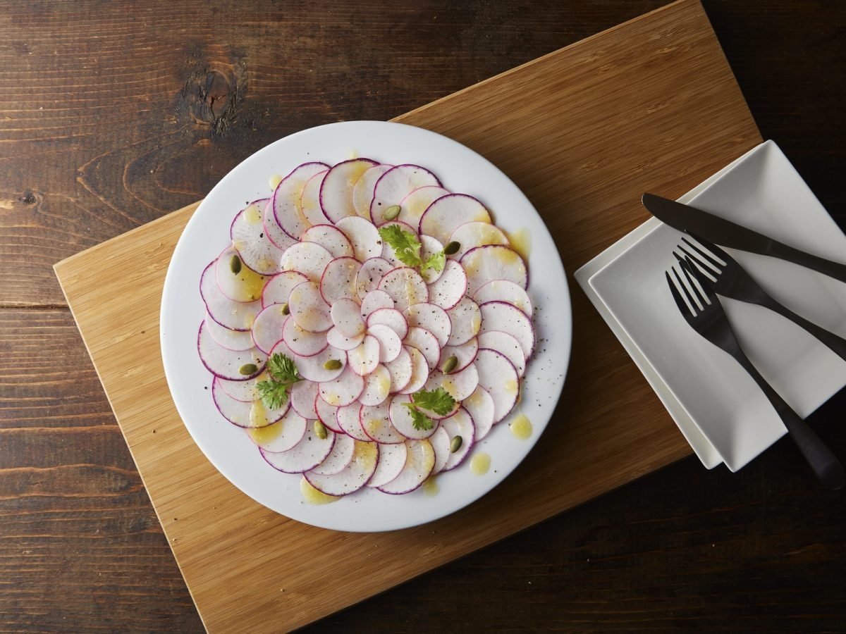 Draufsicht: Ein Kohlrabi-Carpaccio auf einem runden Teller. Der steht steht auf einem Holzbrett. Daneben liegt Essbesteck.