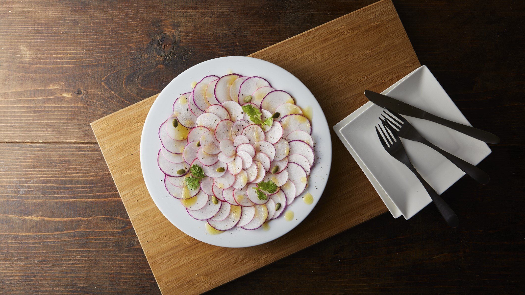 Draufsicht: Ein Kohlrabi-Carpaccio auf einem runden Teller. Der steht steht auf einem Holzbrett. Daneben liegt Essbesteck.