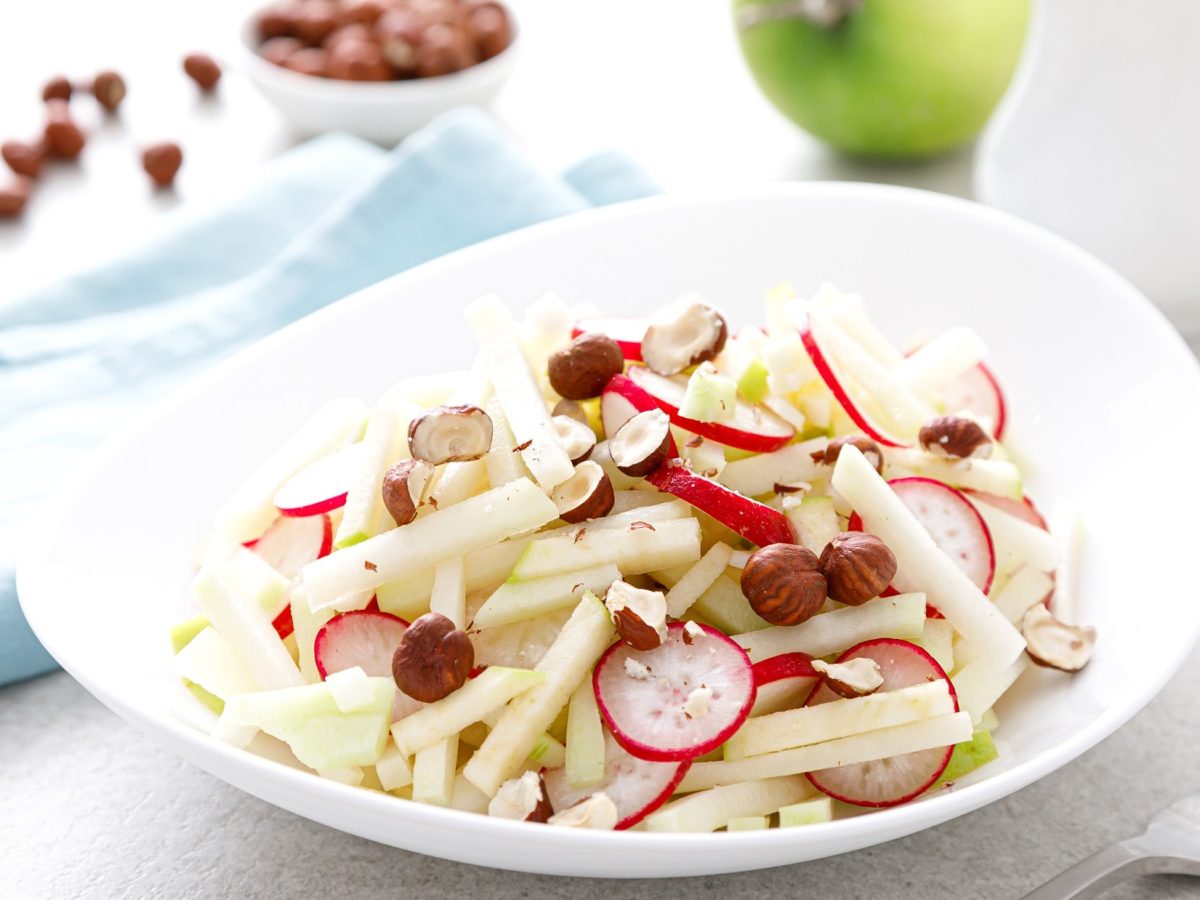 Frontal, leichte Draufsicht: ein Kohlrabi‑Radieschen‑Salat in einem hellen Teller; Drumherum ein Apfel und frische Nusskerne.