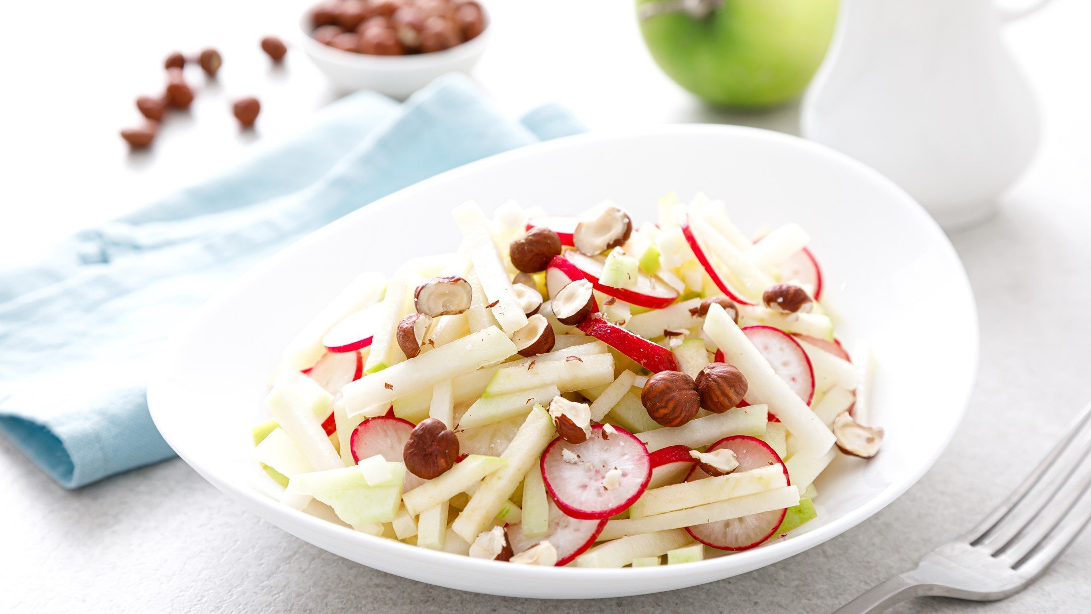 Frontal, leichte Draufsicht: ein Kohlrabi‑Radieschen‑Salat in einem hellen Teller; Drumherum ein Apfel und frische Nusskerne.
