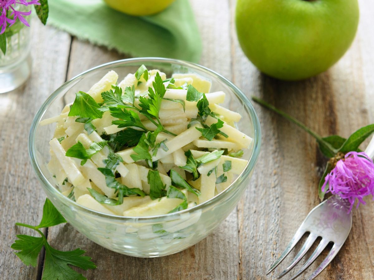 Draufsicht schräg: Ein Kohlrabi‑Rohkostsalat in einer Glasschüssel.. drum herum liegen Äpfel, Blüten und eine Gabel: