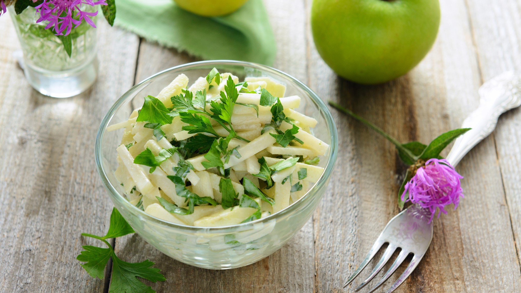 Draufsicht schräg: Ein Kohlrabi‑Rohkostsalat in einer Glasschüssel.. drum herum liegen Äpfel, Blüten und eine Gabel:
