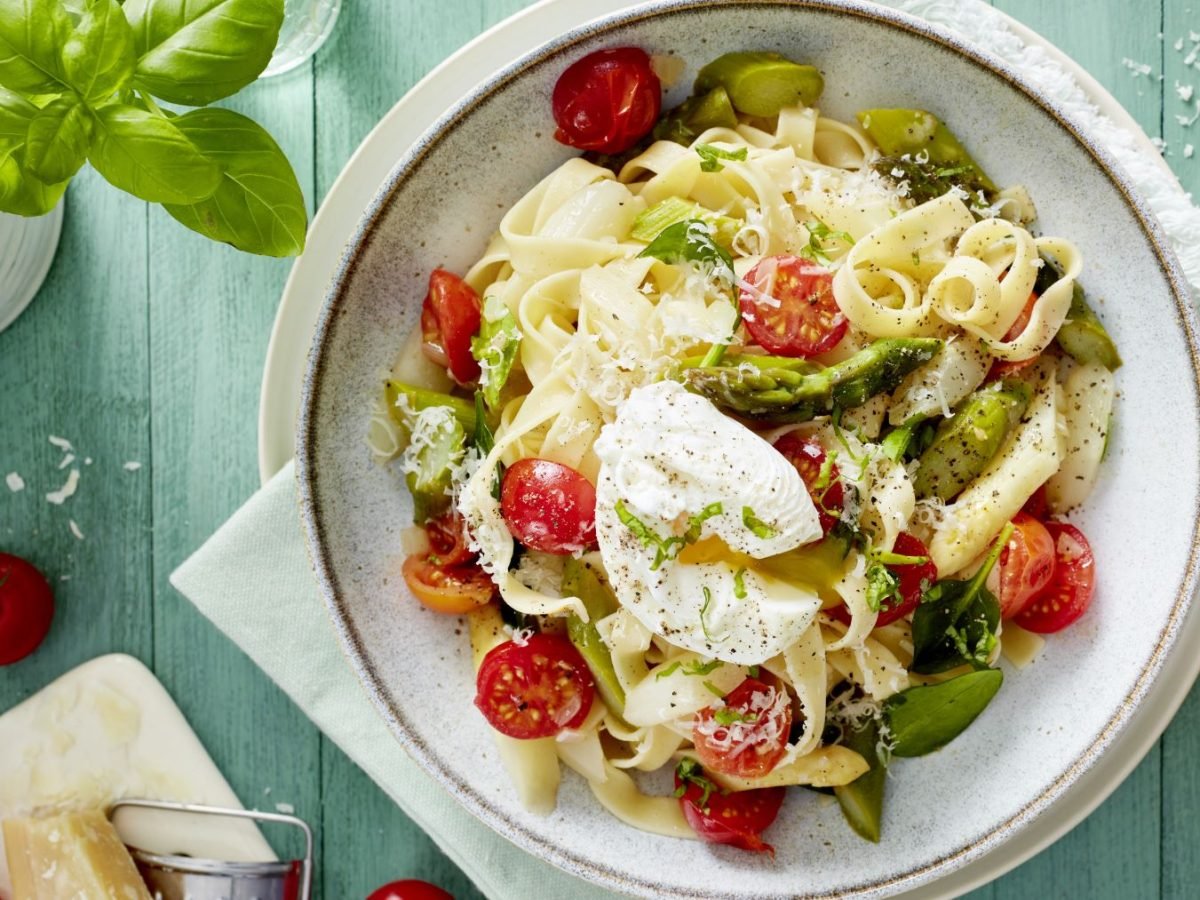 Pasta mit pochiertem Ei in einem weißen Teller angerichtet vor hellgrünem Hintergrund. Links auf dem Foto Basilikum, Tomaten und Parmesan. Rechts ein Schälchen mit Salz.