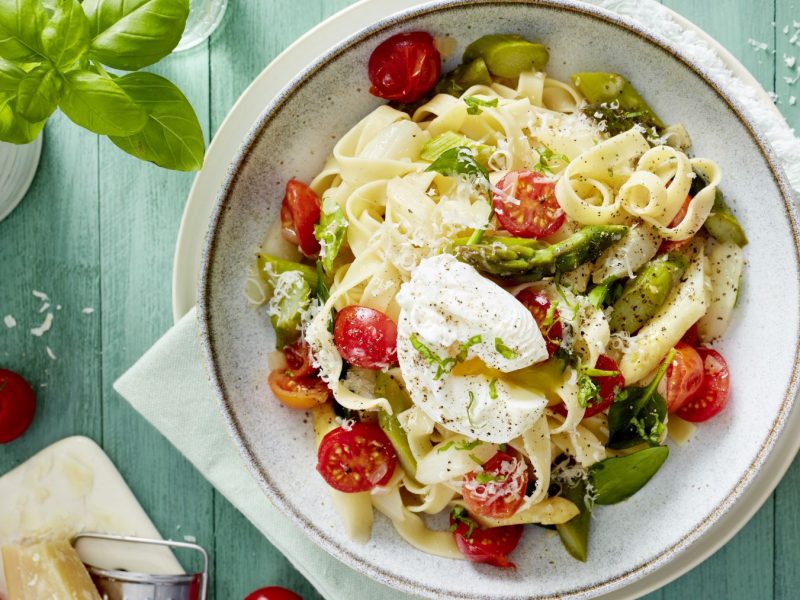 Pasta mit pochiertem Ei in einem weißen Teller angerichtet vor hellgrünem Hintergrund. Links auf dem Foto Basilikum, Tomaten und Parmesan. Rechts ein Schälchen mit Salz.