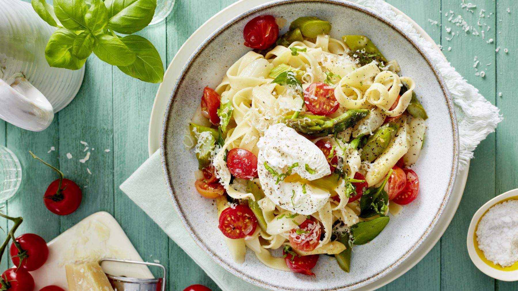 Pasta mit pochiertem Ei in einem weißen Teller angerichtet vor hellgrünem Hintergrund. Links auf dem Foto Basilikum, Tomaten und Parmesan. Rechts ein Schälchen mit Salz.
