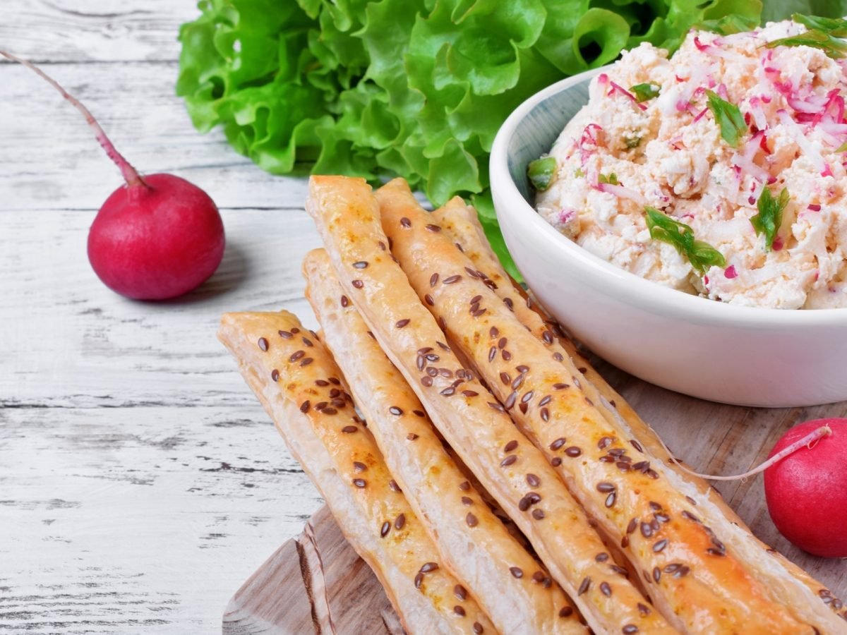 Ein Radieschen-Dip mit Hüttenkäse in einer Schüssel, dünne Brotstangen, frische Radieschen und Salatkopf.