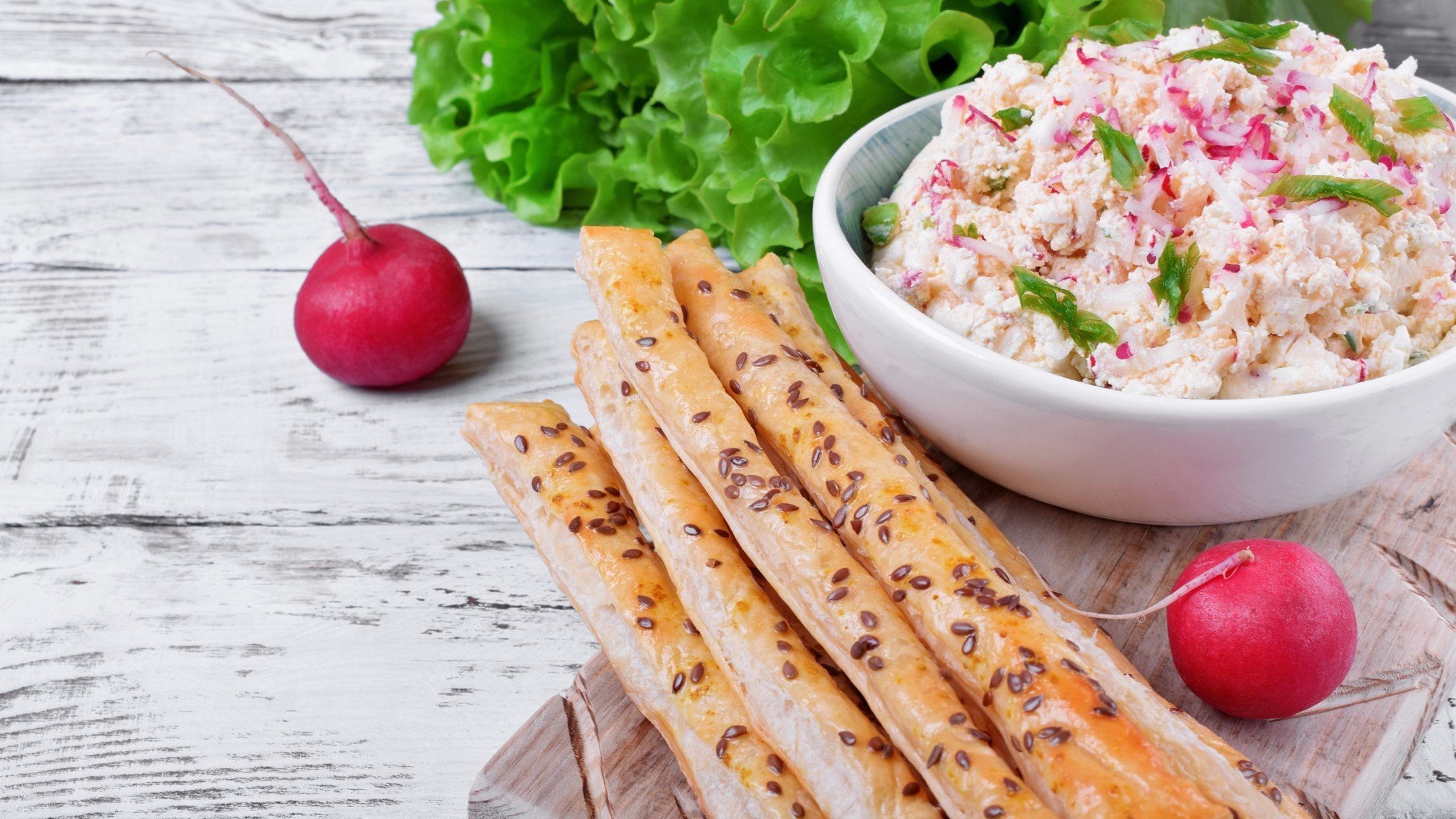 Ein Radieschen-Dip mit Hüttenkäse in einer Schüssel, dünne Brotstangen, frische Radieschen und Salatkopf.