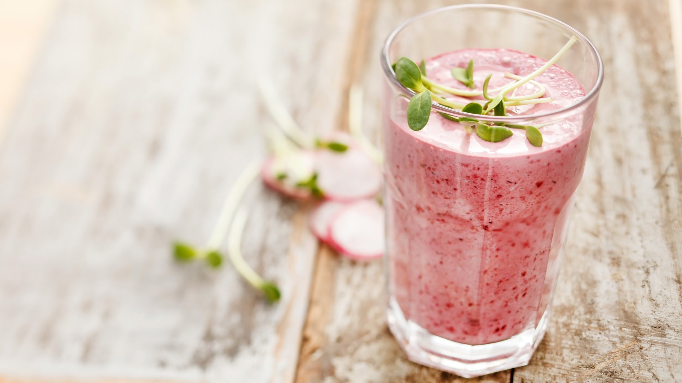 Draufsicht, schräg: Ein Radieschen-Smoothie im Glas auf einem Untergrund aus Holz. Mit frischen Sprossen dekoriert, die auch neben dem Getränk liegen.