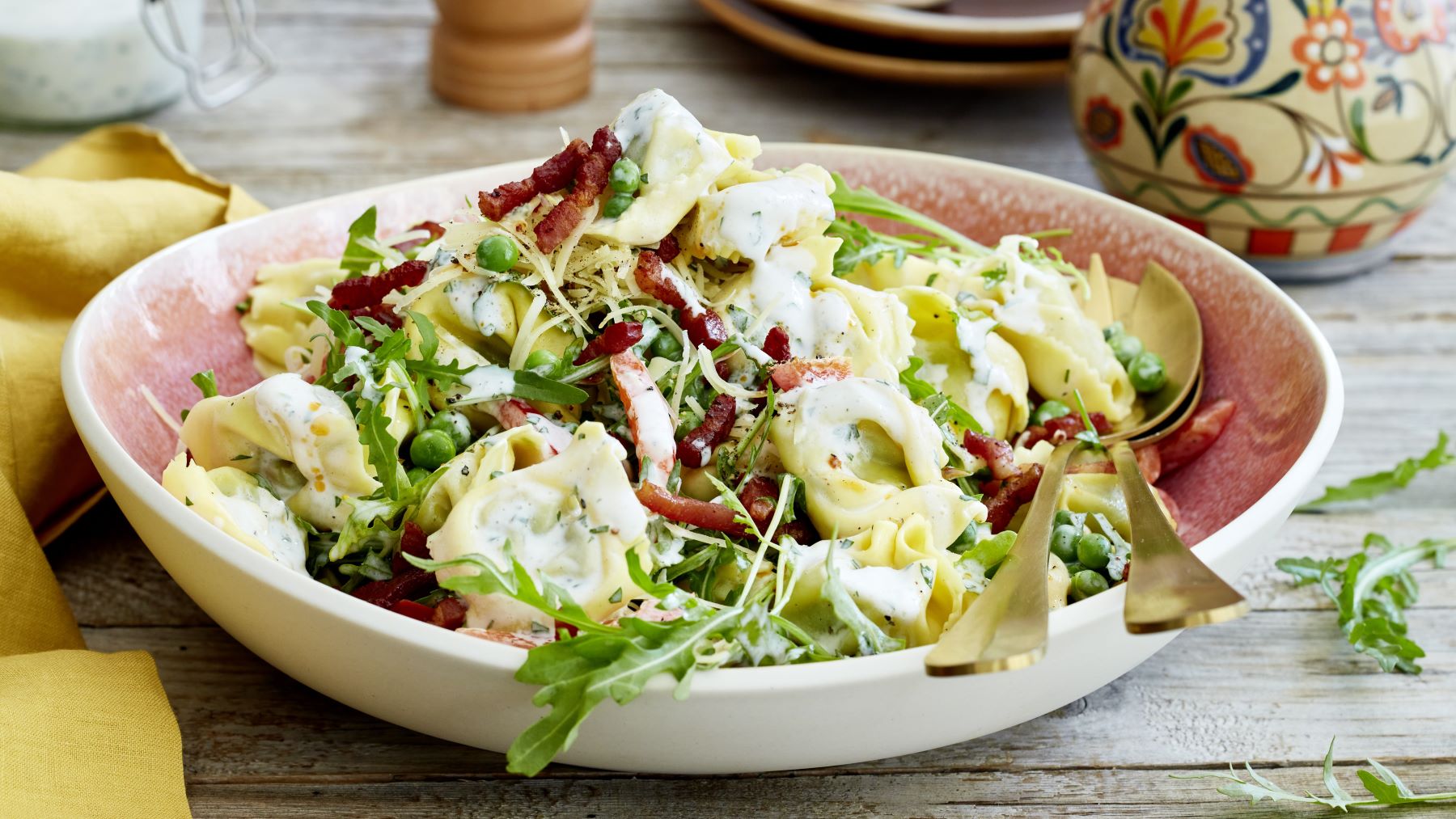 Tortelloni-Salat vor hölzernem Hintergrund. Links ein gelbes Geschirrtuch, rechts eine Vase und eine Pfeffermühle.