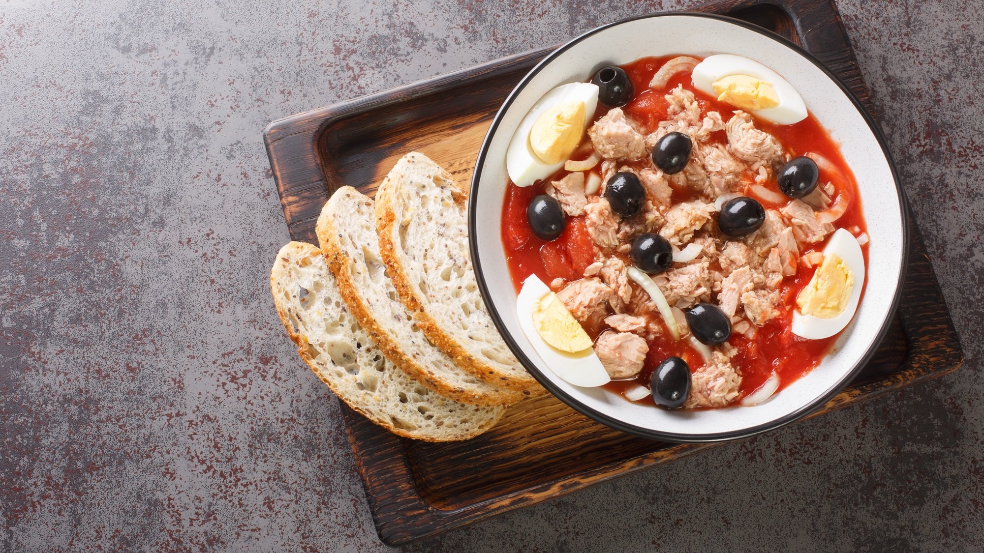Rustikale Servierplatte mit Ensalada Murciana aus Tomaten, Eiern, Oliven und Thunfisch, angerichtet mit Brotscheiben.