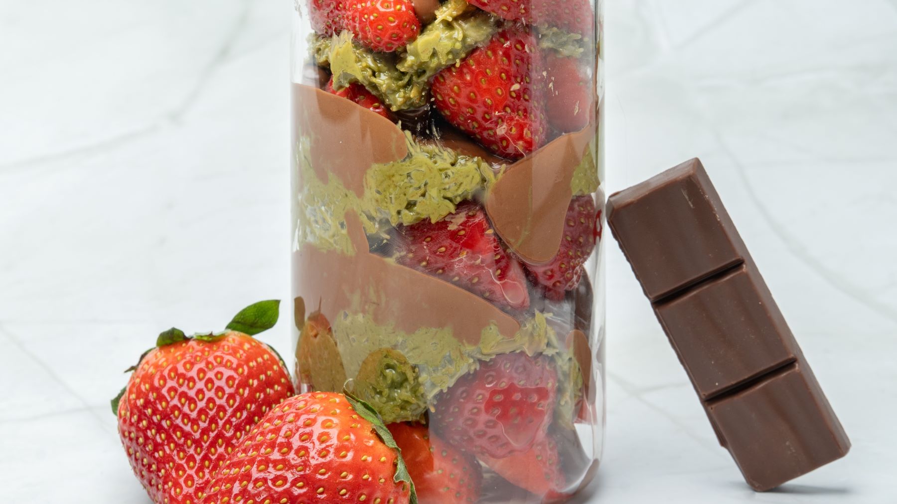 "Erdbeer-Dubai-Becher im Glas, geschichtet mit frischen Erdbeeren, Pistaziencreme, Kokosjoghurt und knackiger Schokoglasur, dekorativ angerichtet mit herablaufender Schokolade.