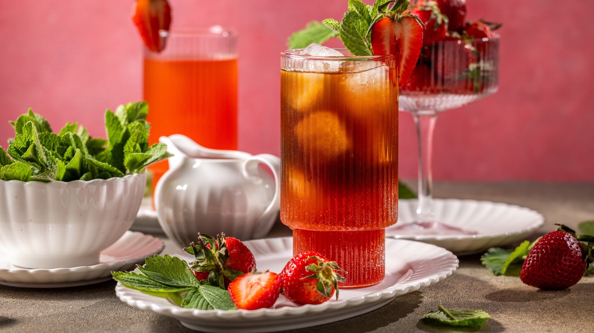 Ein hohes Glas mit eisgekühltem Erdbeer-Eistee, garniert mit frischer Minze und halber Erdbeere, steht auf weißem Porzellanteller.