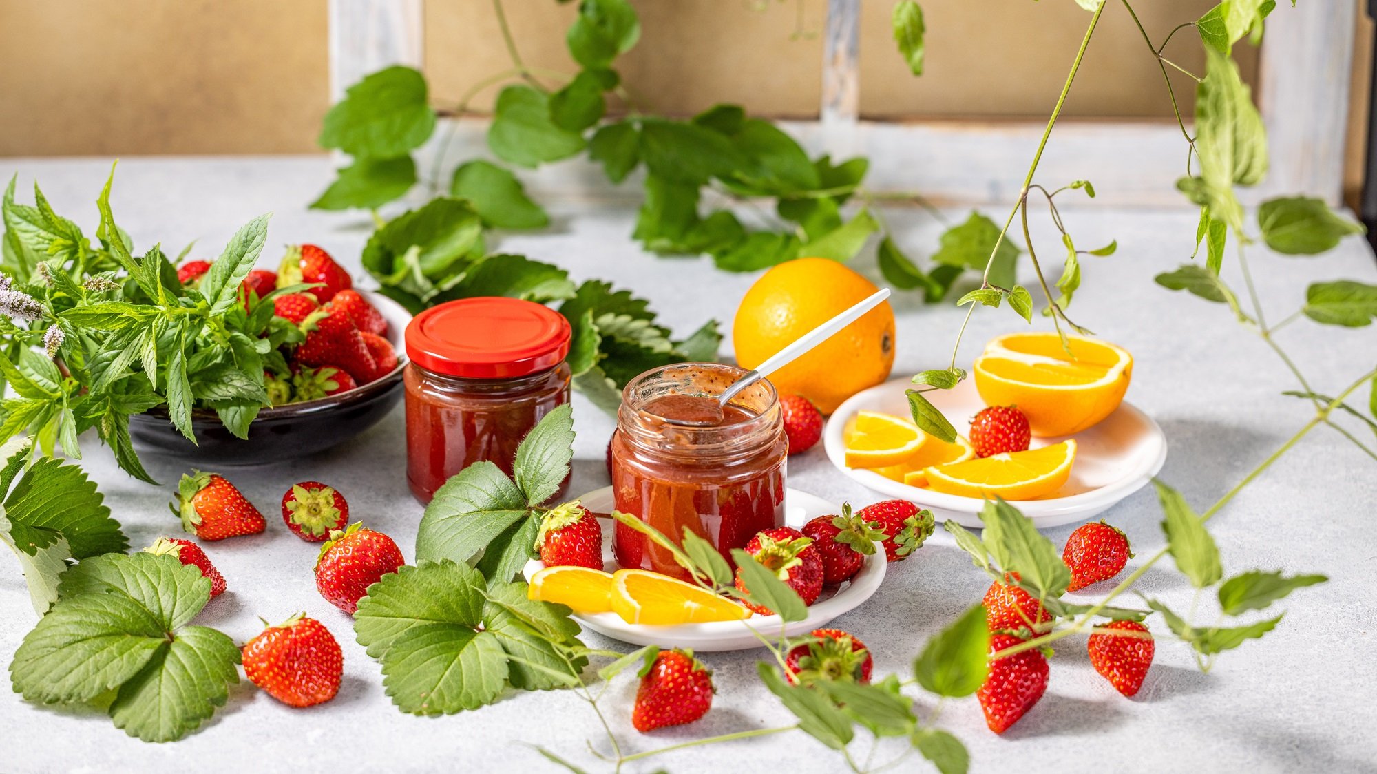 Geöffnete Erdbeer-Orangen-Marmelade mit Löffel steht zwischen frischen Früchten, Minzzweigen und geschnittener Orange auf weißem Teller.