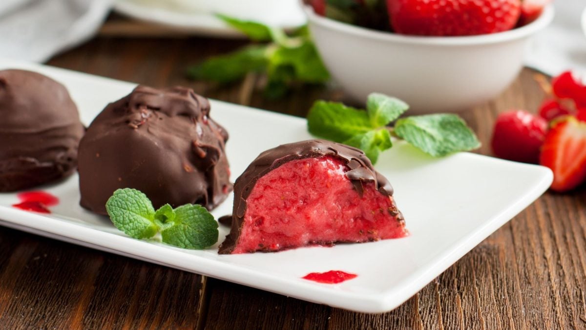 Erdbeer-Schoko-Bites auf einem weißen Teller angerichtet mit Minze vor hölzernem Hintergrund. Hinten auf dem Foto ein Schälchen mit Erdbeeren.