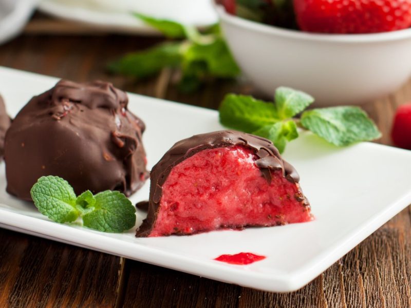 Erdbeer-Schoko-Bites auf einem weißen Teller angerichtet mit Minze vor hölzernem Hintergrund. Hinten auf dem Foto ein Schälchen mit Erdbeeren.