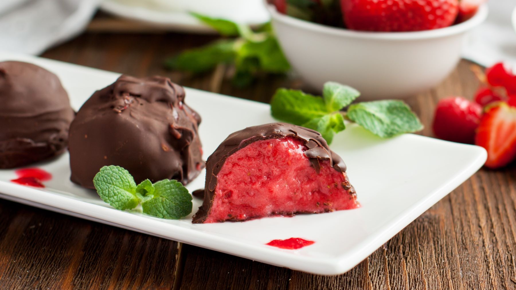 Erdbeer-Schoko-Bites auf einem weißen Teller angerichtet mit Minze vor hölzernem Hintergrund. Hinten auf dem Foto ein Schälchen mit Erdbeeren.
