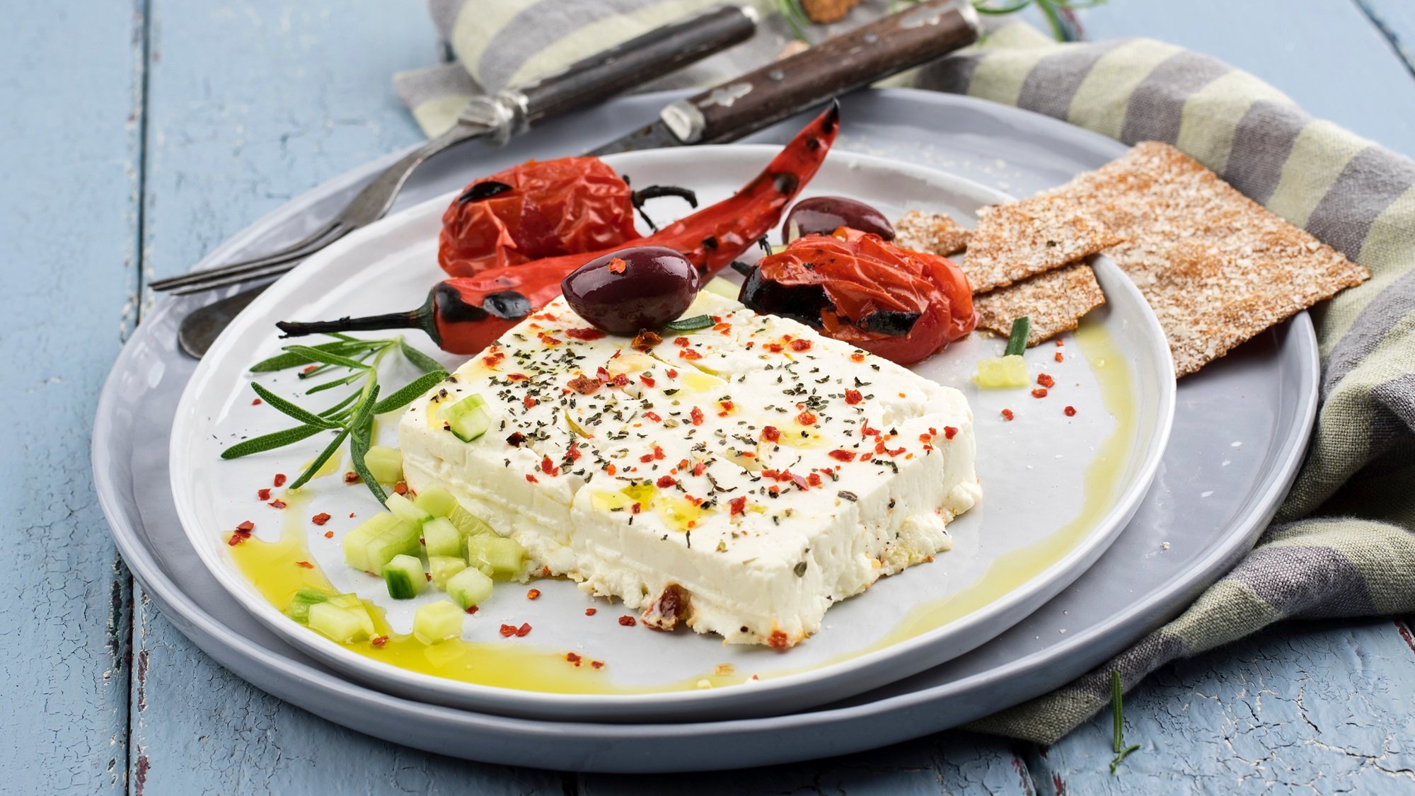 Draufsicht auf ein Stück gegrillten Feta, de rmit Kärutern, Grillgemüse, Gurke und einer Olive garniert ist.