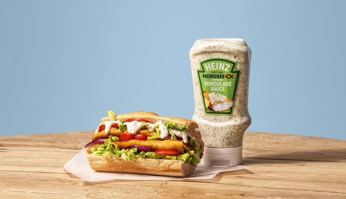 Flasche der veganen Remoulade von Heinz und Nordsee vor einem blauen Hintergrund und auf einem Holzbrett, daneben üppig belegtes Fisch-Sandwich.