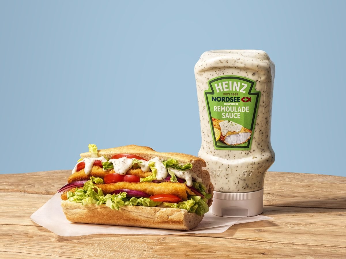 Flasche der veganen Remoulade von Heinz und Nordsee vor einem blauen Hintergrund und auf einem Holzbrett, daneben üppig belegtes Fisch-Sandwich.