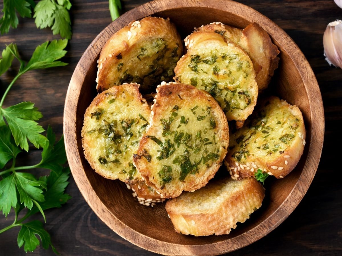 Knusprige Knoblauchbrote mit Kräutern und Sesam in Holzschale, daneben frische Petersilie und Knoblauchzehen auf dunklem Tisch.