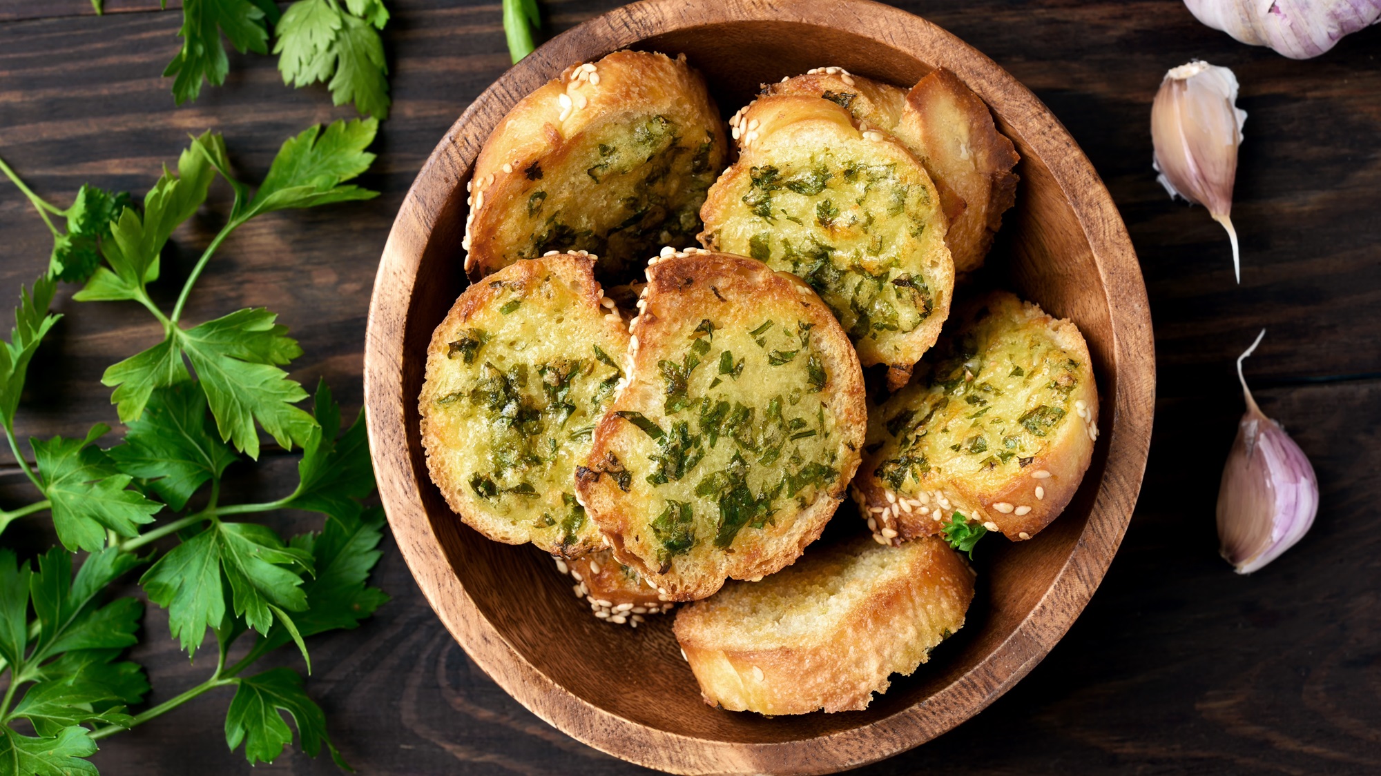 Knusprige Knoblauchbrote mit Kräutern und Sesam in Holzschale, daneben frische Petersilie und Knoblauchzehen auf dunklem Tisch.