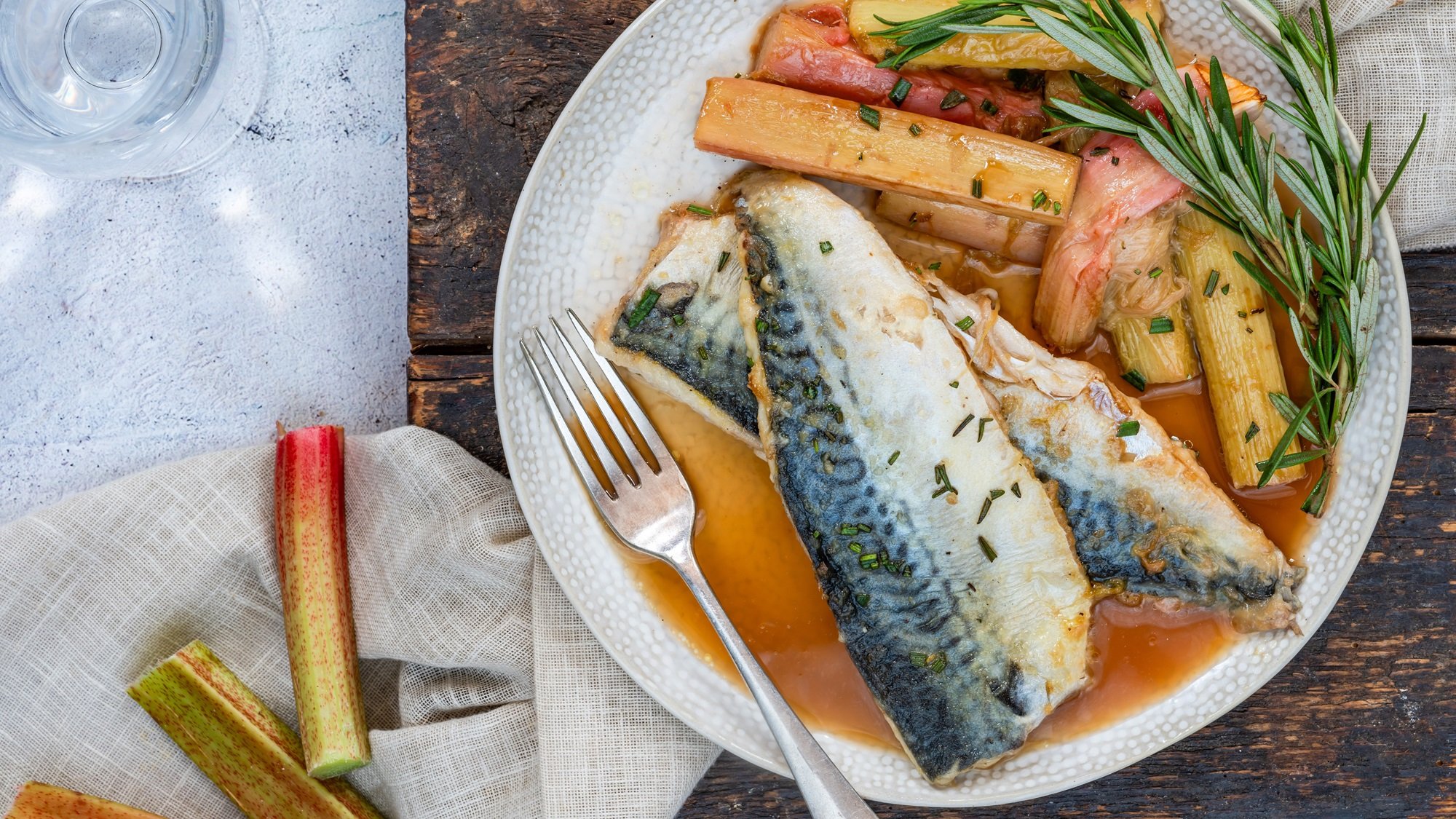 Nahaufnahme eines Fischgerichts mit saftigem Makrelenfilet, Rhabarber, Kräutern und hellem Leinentuch auf Holzplatte.