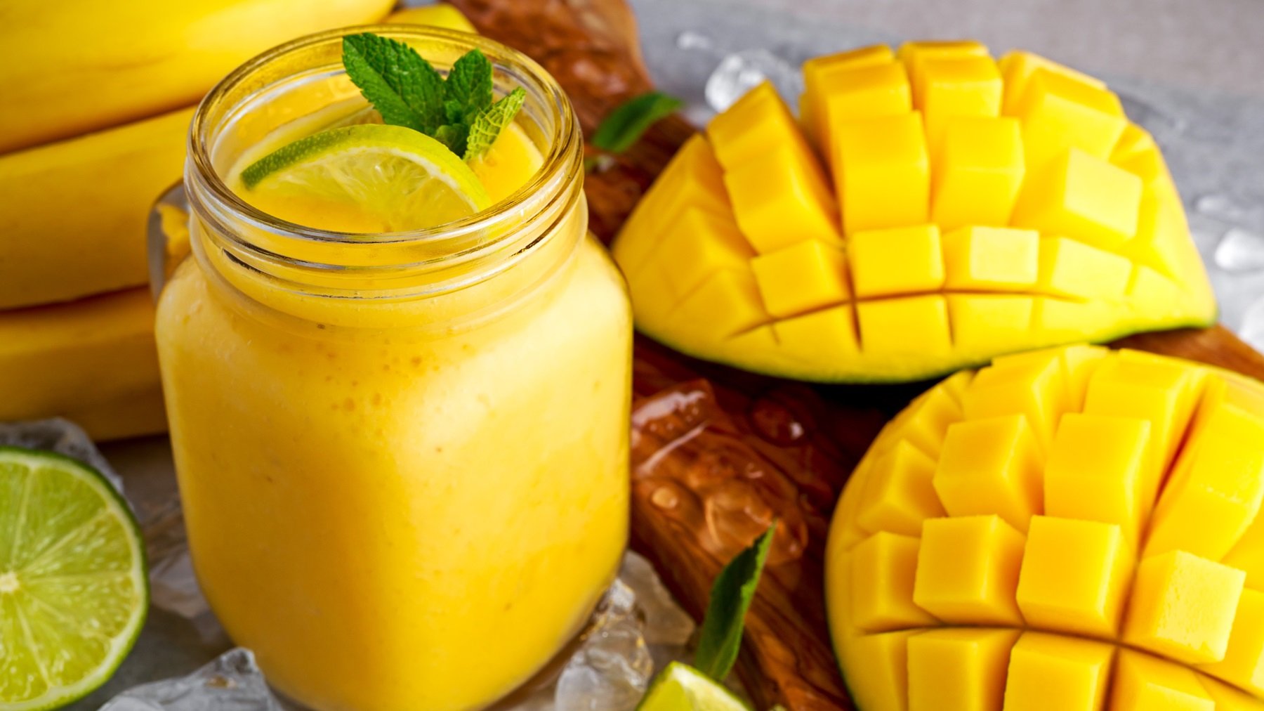 Ein Glas mit dem gelben Smoothie sowie frischer Mango und Banane im Hintergrund. Alles von schräg oben auf grauem Untergrund fotografiert.