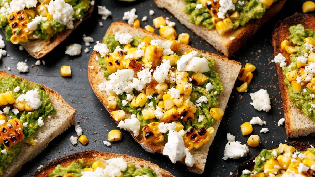 Nahaufnahme von knusprigen Avocado-Stullen mit gegrilltem Mais und Feta, dazwischen verstreute Krümel auf schwarzem Untergrund.