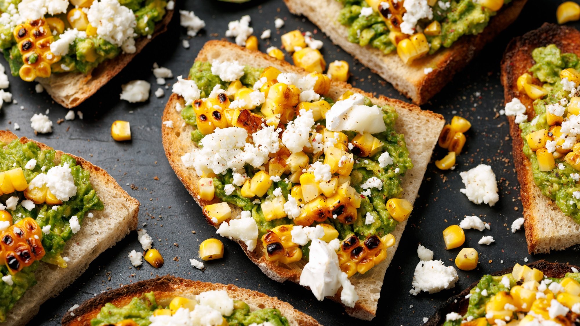 Nahaufnahme von knusprigen Avocado-Stullen mit gegrilltem Mais und Feta, dazwischen verstreute Krümel auf schwarzem Untergrund.