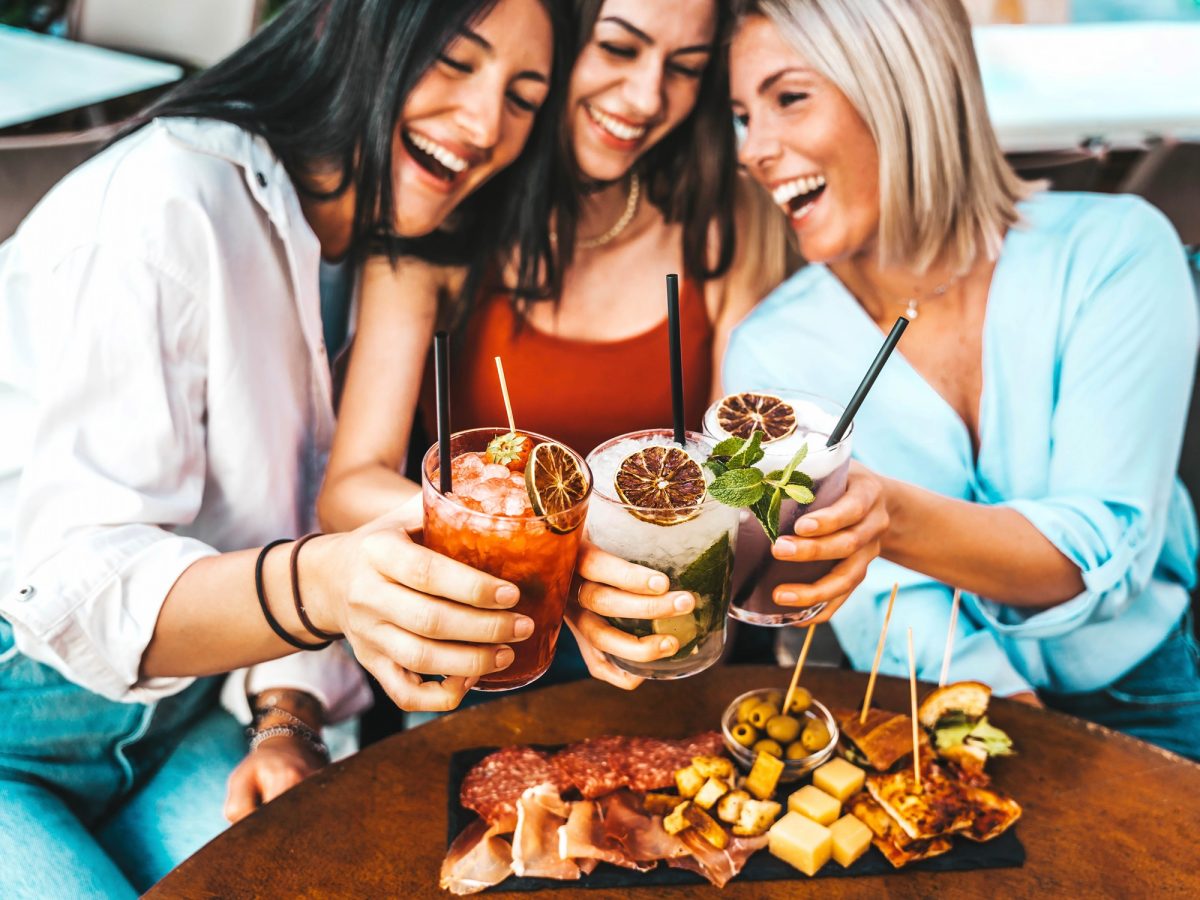 Fröhliche Runde junger Frauen mit offenen Haaren und farbiger Kleidung stoßen lachend mit Mocktails an, vor ihnen mediterrane Snacks.