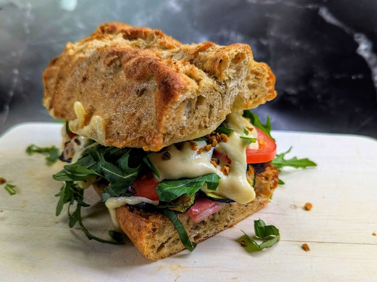 Rustikales Sandwich mit dicker Kruste, gefüllt mit Mortadella, Tomate, Rucola und geschmolzener Parmesancreme auf Holzbrett.