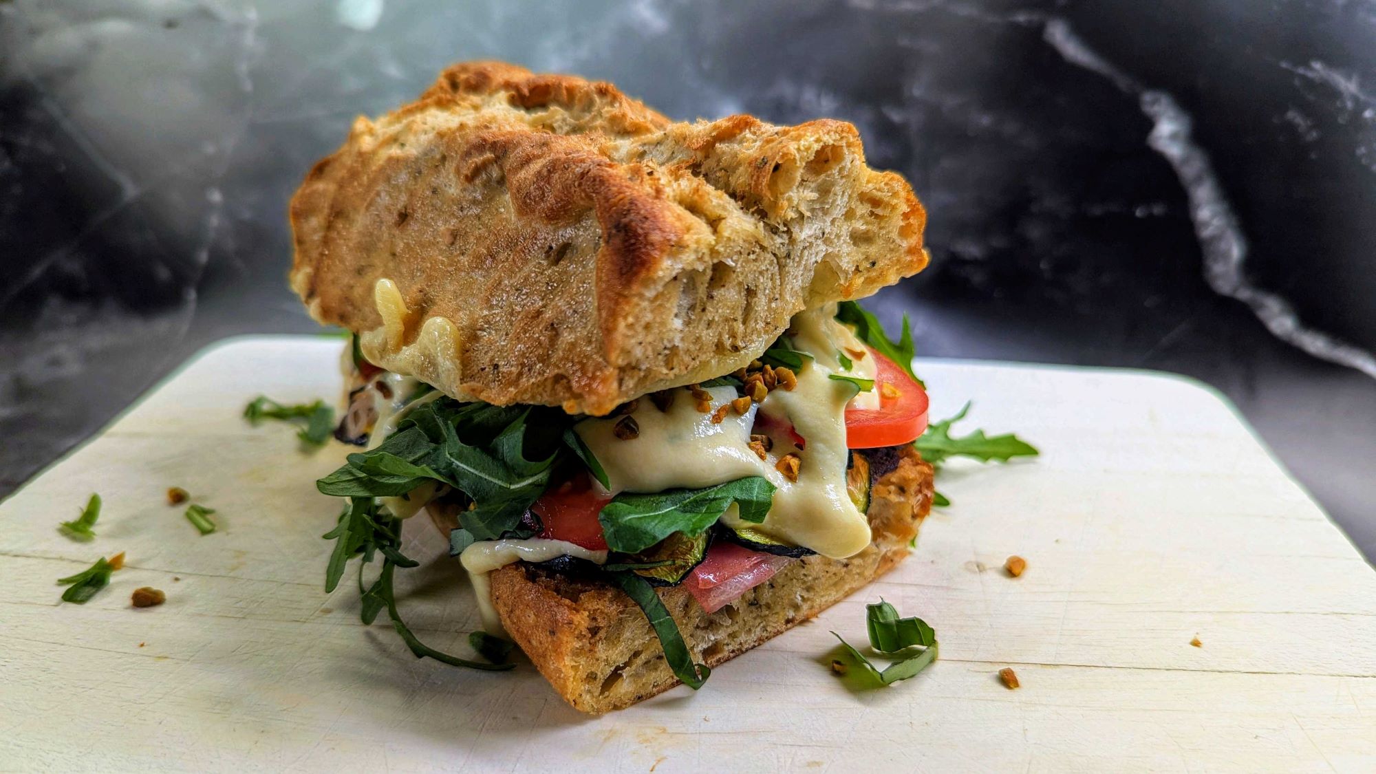 Rustikales Sandwich mit dicker Kruste, gefüllt mit Mortadella, Tomate, Rucola und geschmolzener Parmesancreme auf Holzbrett.
