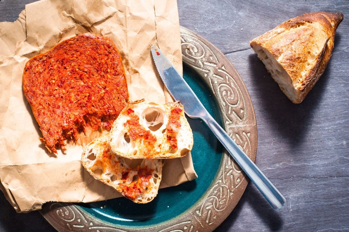 Zwei Scheiben Weißbrot mit würziger ’Nduja auf braunem Papier neben einem Messer auf grünem Keramikteller angerichtet.