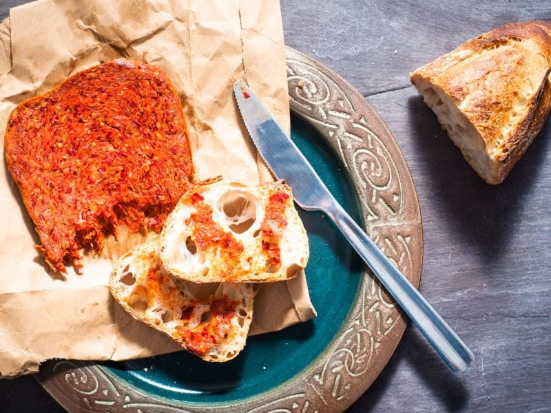 Zwei Scheiben Weißbrot mit würziger ’Nduja auf braunem Papier neben einem Messer auf grünem Keramikteller angerichtet.