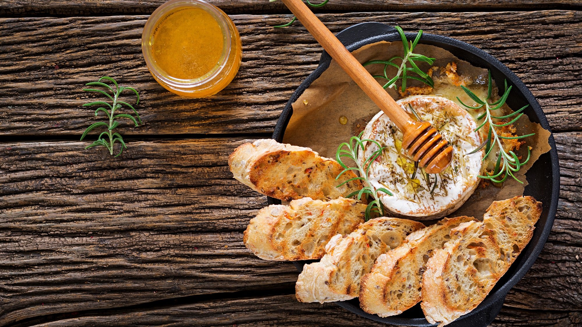 Ofenkäse mit Rosmarin und Honig in schwarzer Pfanne, serviert mit knusprigen Baguettescheiben auf rustikalem Holzuntergrund.