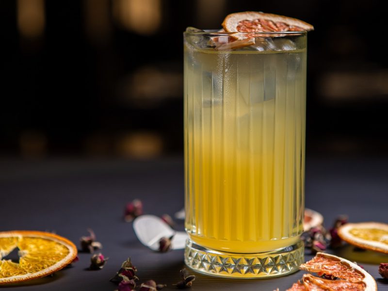 Ein Glas mit dem Padovani Spritz auf einem dunklen Tisch, mit Blüten und getrockneten Orangenscheiben. Alles von der Seite fotografiert.