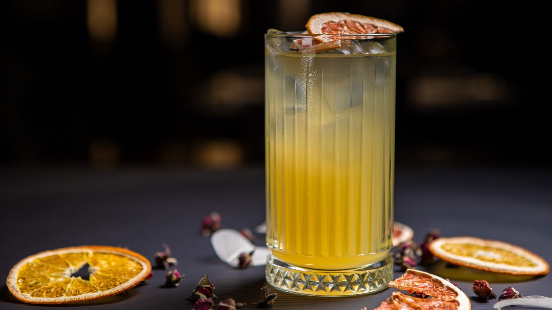 Ein Glas mit dem Padovani Spritz auf einem dunklen Tisch, mit Blüten und getrockneten Orangenscheiben. Alles von der Seite fotografiert.