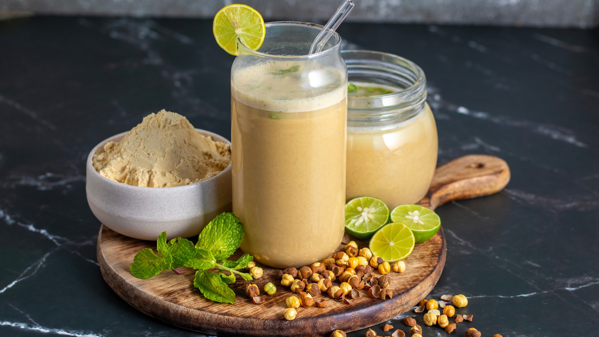 Ein Glas Sattu-Getränk mit Limettenscheibe, daneben Schale mit Mehl, Minze, halbierte Limetten und geröstete Kichererbsen.