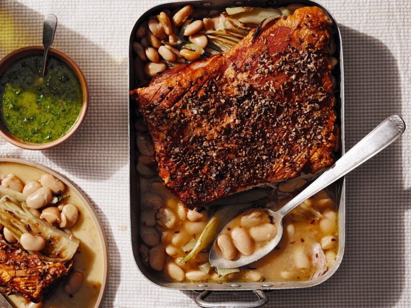 Eine Auflaufform mit dem Schweinebauch und daneben eine kleine grüne Schale mit dem Dip. Alles im Sonnenlicht direkt von oben fotografiert auf heller Tischdecke.