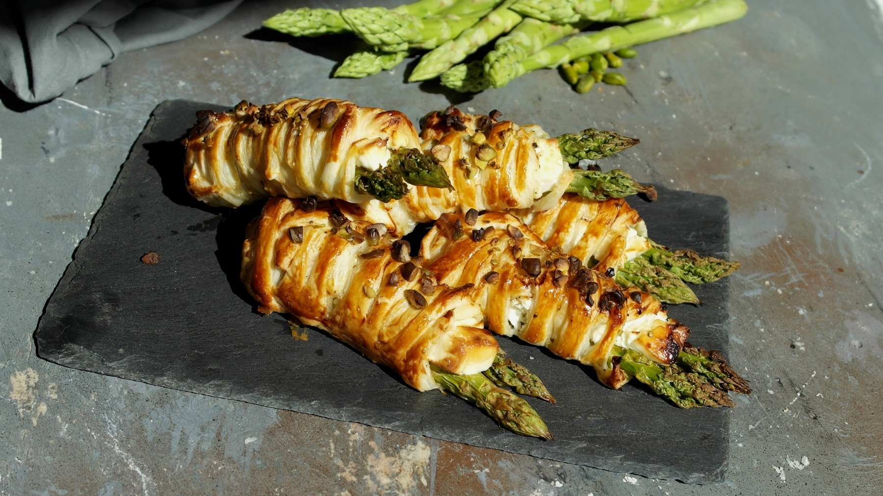 Eine schwarze Schieferplatte mit dem Gebäck auf einem grauen Steintisch mit grünem Spargel im Hintergrund. Alles im Sonnenlicht fotografiert.