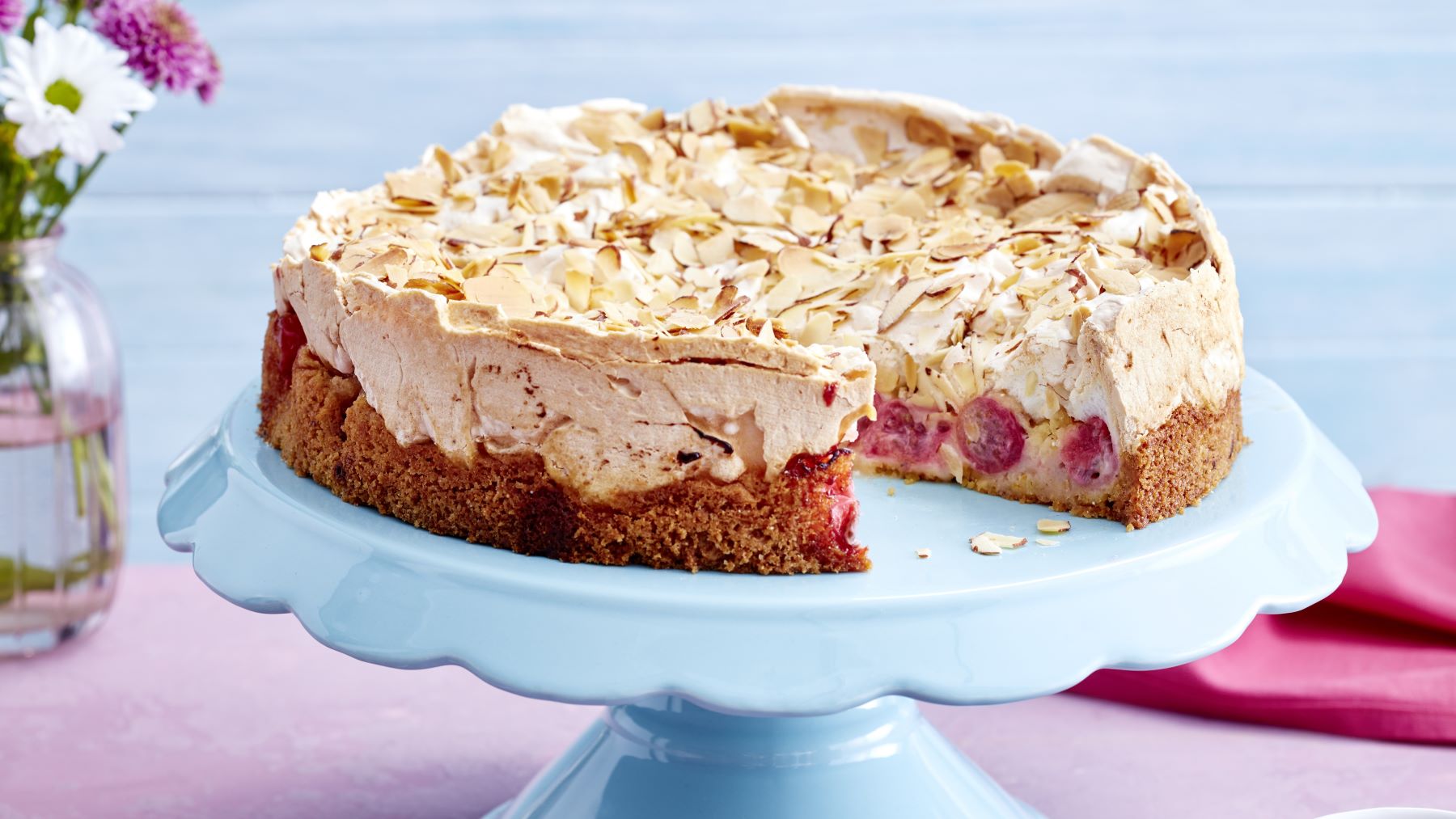 Stachelbeer-Baiser-Kuchen auf einem hellblauen Tortenteller vor rosa und blauem Hintergrund. Links Blumen und rechts ein pinkes Geschirrtuch.