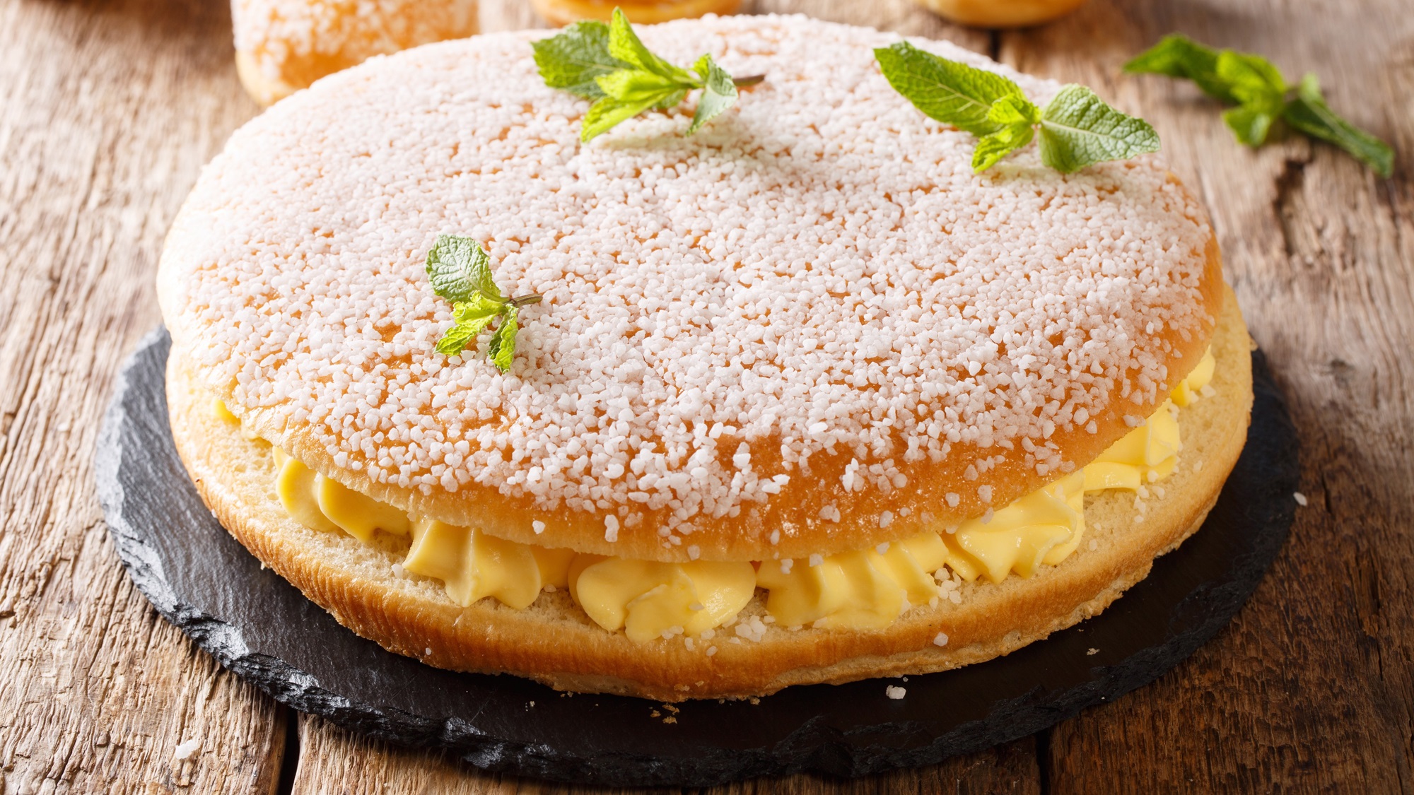 Französische Tarte Tropézienne mit knuspriger Zuckerkruste, üppiger Vanillecreme und Minzblättern auf rustikalem Holztisch.