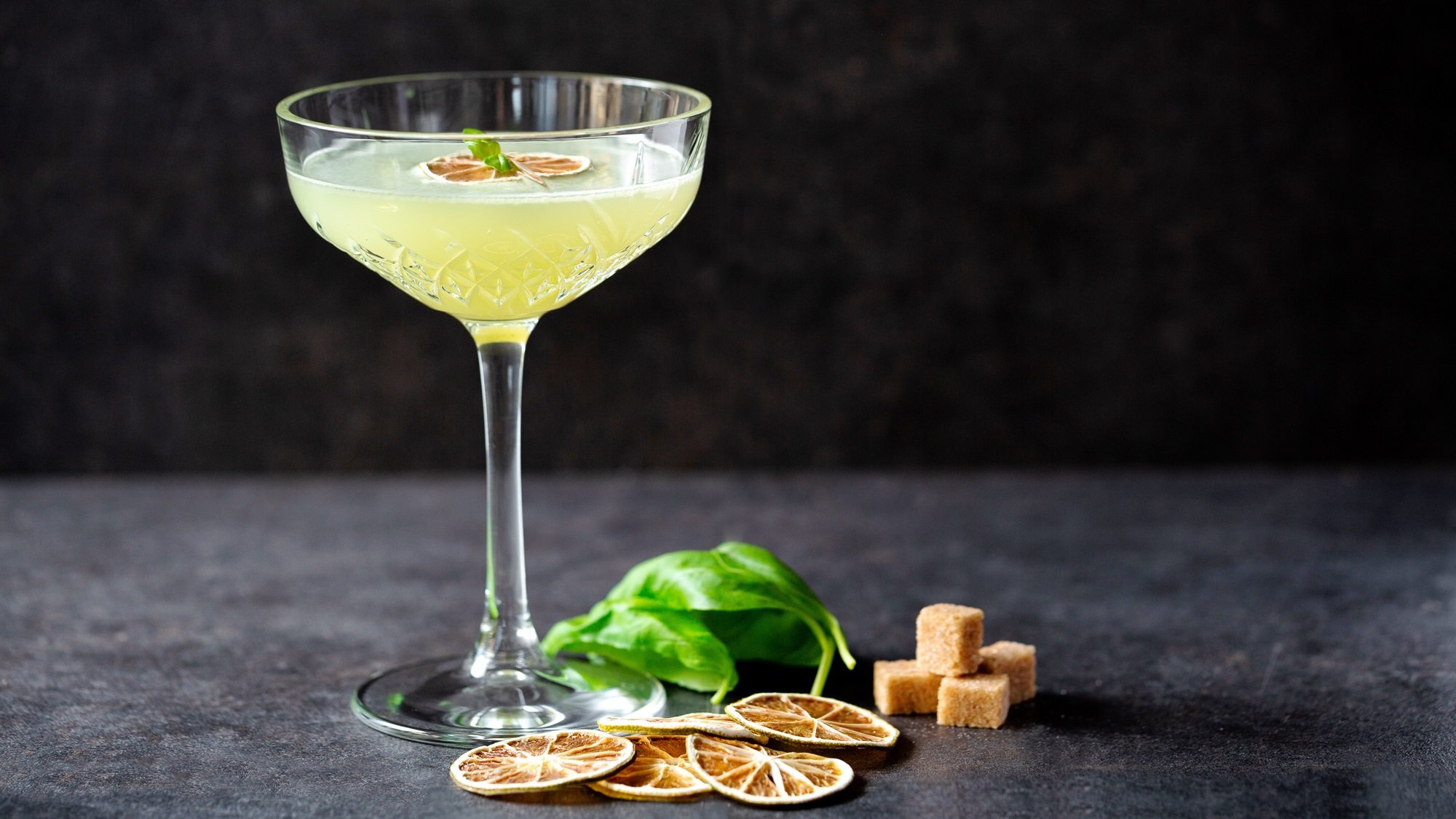 Kristallklares Glas mit spritzigem Thai-Basilikum Daiquiri, dekoriert mit grünen Basilikumblättern und braunen Zuckerwürfeln.